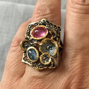 Peut inclure: Une bague en or et argent avec un sertissage de pierres précieuses roses et bleues. La bague présente un design délicat et complexe avec des motifs floraux.
