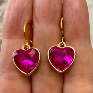 Peut inclure: Une paire de boucles d'oreilles dorées, chacune ornée d'une pierre précieuse fuchsia en forme de cœur. Les pierres précieuses sont serties dans un cadre doré, et les boucles d'oreilles ont une fermeture à levier.