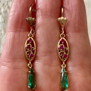 Peut inclure: Boucles d'oreilles pendantes dorées avec des rubis et des émeraudes vertes en forme de goutte. Elles présentent un design ovale avec des grappes de rubis et des détails de filigrane, pour un style vintage.