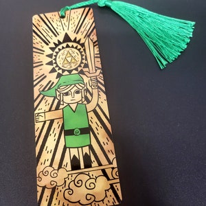 May include: A gold and green bookmark featuring a cartoon illustration of Link from the Legend of Zelda video game series. Link is holding a sword and standing on clouds with a golden sun and a Triforce symbol behind him. The bookmark has a green tassel.
