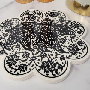 May include: A black and white floral design trivet with a scalloped edge. The trivet is made of ceramic and is perfect for protecting surfaces from hot dishes.