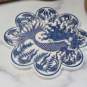 May include: Blue and white ceramic trivet with a mermaid design. The mermaid is surrounded by seaweed and seashells.