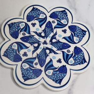 May include: A white ceramic trivet with a blue and white fish design. The trivet is shaped like a flower with 10 petals.