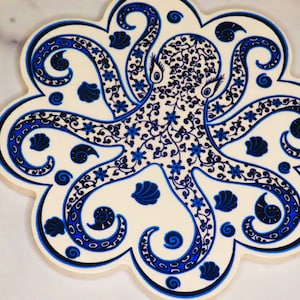 May include: A cream-colored, octagonal ceramic trivet featuring a blue octopus design. The octopus has intricate floral and swirl patterns, with seashell and spiral accents. The trivet is approximately 8 inches in diameter.
