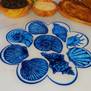 May include: A white ceramic trivet with a blue and white seashell design. The trivet is shaped like a flower with eight petals.