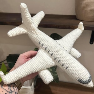 May include: A handmade, white crochet airplane toy with gray accents. The toy features wings, a tail, and a detailed design, perfect for children's play or nursery decor. The airplane is held in a person's hand.