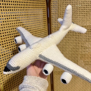 May include: A handcrafted crochet aeroplane toy made with off-white yarn. The aircraft features four engine pods with black circular centres, a grey cockpit window detail, and grey stripes along the wings. The structure is soft and textured.