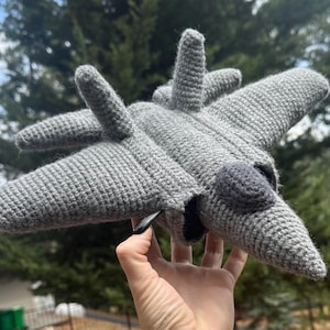 Könnte beinhalten: Ein handgefertigtes graues Häkel-Spielzeugflugzeug mit Flügeln, Heck und Cockpit-Details. Das Spielzeug wird in einer Hand vor einem verschwommenen grünen Hintergrund aus Bäumen gehalten. Das Flugzeug ist ein weiches, strukturiertes Bastelobjekt.