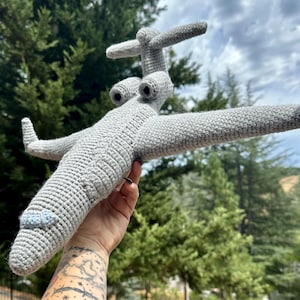 Könnte beinhalten: Ein handgefertigtes, graues gehäkeltes Spielzeugflugzeug mit hellblauer Nase. Das Spielzeug hat detaillierte Flügel, ein Heck und zwei Strahltriebwerke. Das Flugzeug wird vor dem Hintergrund grüner Bäume und eines bewölkten Himmels gehalten.