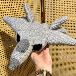 Puede incluir: Un juguete de avión tejido a mano en gris con detalles en negro. El juguete presenta un diseño detallado con alas, cabina y aletas de cola. El hilo suave y texturizado le da un aspecto único y artesanal. Un artículo decorativo divertido.