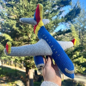 Puede incluir: Un avión de juguete de ganchillo hecho a mano. El cuerpo es azul con alas y cola grises. La cola presenta detalles rojos, amarillos y azules. El juguete se sostiene sobre un fondo de follaje verde y cielo azul.
