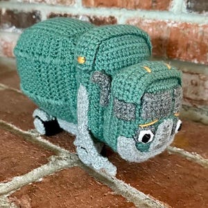 May include: A handmade crochet toy truck in shades of teal, gray, and black. The truck features a detailed design with a cab, bed, and wheels. The toy is crafted with a textured stitch, giving it a soft, plush appearance. The truck is a charming, handcrafted item.