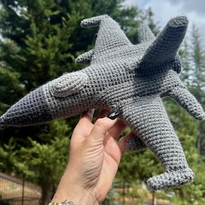 Puede incluir: Un juguete de avión gris tejido a ganchillo con una nariz degradada. El juguete hecho a mano presenta alas, una cola y otros detalles. El avión se sostiene frente a un fondo verde borroso.