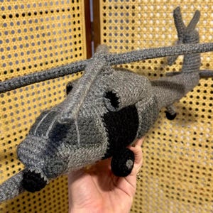 Puede incluir: Un helicóptero de ganchillo hecho a mano en tonos grises y negros. El helicóptero de juguete presenta un diseño detallado con rotores, cola y cabina. El trabajo de ganchillo es visible, mostrando la textura y la artesanía del artículo.