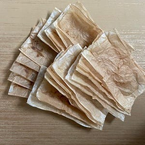 May include: A stack of square, light brown paper sheets with a textured, crinkled appearance. The paper has a slightly translucent quality and varying shades of brown, suggesting a natural or aged aesthetic. The sheets are arranged on a light brown wooden surface.