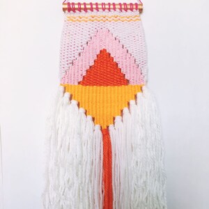 May include: A woven wall hanging with a geometric pattern in white, pink, orange, and yellow. The top has a copper rod with pink and orange details. White yarn tassels hang from the bottom, adding texture. The piece is a decorative item.