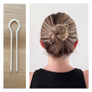 May include: Silver hair stick with a curved design. The stick is being used to hold a bun in place.