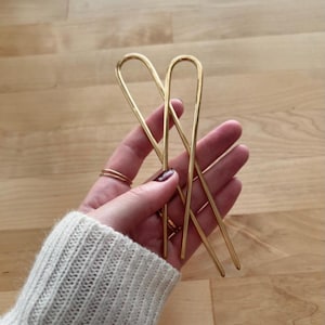 May include: Two gold hair pins with a curved end. The pins are held in a hand.
