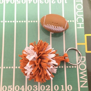 May include: A white and orange pom-pom keychain with a silver key ring. The pom-pom is made of faux fur and is attached to a brown leather strap. The keychain is sitting on a green and white football field.