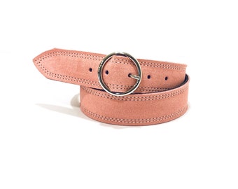May include: A blush pink suede belt with a silver-colored circular buckle. The belt features a stitched design along the edges and is displayed against a white background. The belt is approximately 1.25 inches wide.