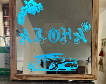 Op de afbeelding: Een houten spiegel met een blauw en wit ontwerp. De spiegel heeft een blauwe "Aloha" inscriptie, een blauwe hibiscusbloem en een blauw silhouet van een auto met een surfboard erop.