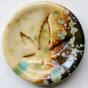 May include: A circular ceramic dish with a leaf-shaped cutout. The dish has a cream base with green and blue speckles, fading to brown and black on the right. The diameter of the dish is approximately 15 cm.