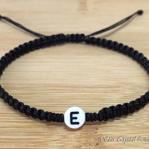 May include: Black macrame bracelet with a white bead featuring the letter "E". The adjustable bracelet is displayed on a light brown wooden surface. The bracelet is a simple, yet stylish accessory.