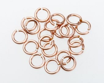 May include: A collection of open copper jump rings, scattered on a white surface. The rings are circular with a small gap, ready for use in jewelry making or other crafts. The copper color provides a warm, metallic aesthetic.