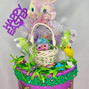 May include: A decorative Easter-themed display featuring a purple base with bunny and egg illustrations. A woven basket holds two plush birds. The display is topped with a bunny head, the words "Happy Easter", and decorative grass and flowers.