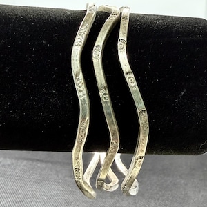 May include: Three silver-toned metal bangles with a wavy, serpentine shape. Each bangle features a series of small, stamped sun-like motifs along the outer surface. The metal has a slightly weathered, matte finish with visible texture throughout.