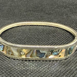 May include: A silver-toned hinged bangle bracelet featuring an inlaid band of iridescent abalone shell. The shell displays swirling patterns of blue, green, and cream colors, accented by small circular silver metal studs embedded within the inlay.