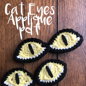 Puede incluir: Cuatro aplicaciones de ojo de gato de ganchillo en amarillo, negro y blanco. Los ojos son de forma ovalada con pupilas negras y reflejos blancos. El texto "Cat Eyes Applique PDF" está escrito encima de los ojos.