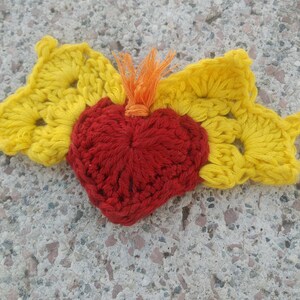 Puede incluir: Un adorno de corazón tejido a crochet con un centro rojo y alas amarillas. El corazón está hecho de hilo rojo y las alas de hilo amarillo.