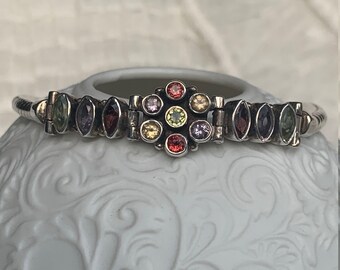 May include: A silver bracelet featuring a central cluster of colourful gemstones, with smaller, oval-shaped gems in various colours. The bracelet has a textured, ribbed design.