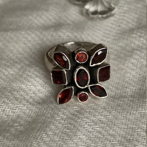Könnte beinhalten: Ein silberner Ring mit einem floralen Design aus dunkelroten Edelsteinen. Der Ring hat ein silbernes Band und ist mit ovalen, quadratischen und runden roten Steinen besetzt. Präsentiert auf hellem, grauem Stoff.