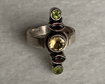 May include: A silver ring featuring a central, round, yellow gemstone, flanked by two smaller, oval, dark red gemstones, and topped and bottomed with green, round gemstones. The ring has a simple band.