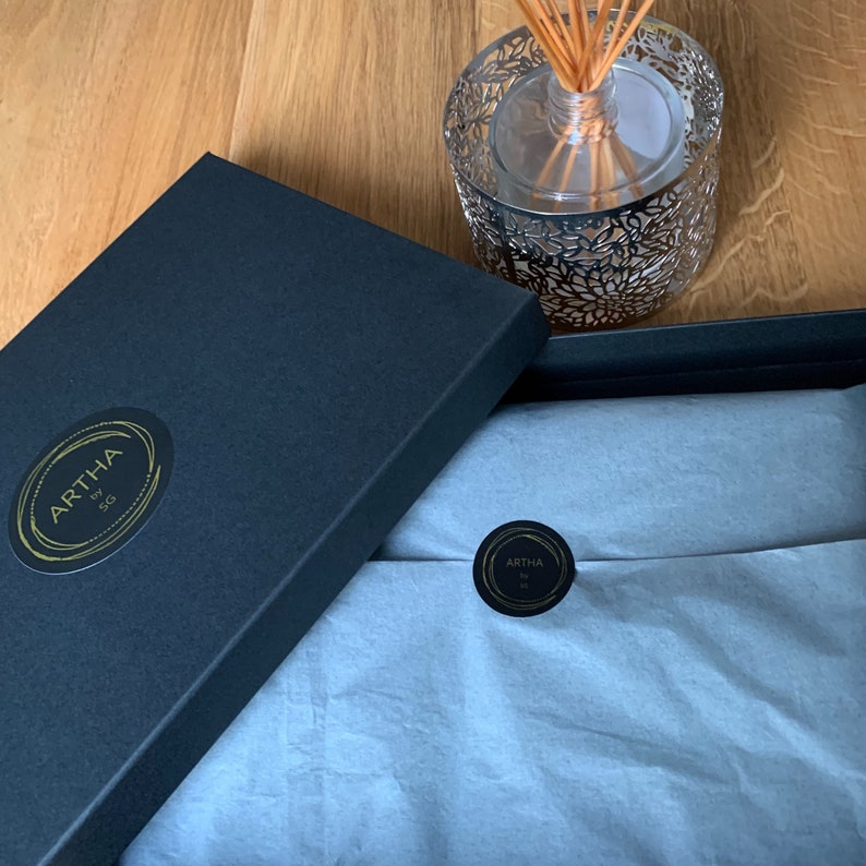 Op de afbeelding: Een zwarte geschenkdoos met een gouden sticker met de tekst "ARTHA by SG". Binnenin lichtblauw vloeipapier. Een zilveren diffuser met stokjes en een kleine sticker zijn ook zichtbaar.