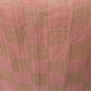 Op de afbeelding: Close-up van een textiel met een geruit patroon. Het patroon wisselt af tussen stoffige roze en bruine vierkanten. De stof heeft een licht getextureerd uiterlijk, wat wijst op een geweven materiaal. De algehele esthetiek is zacht en neutraal.