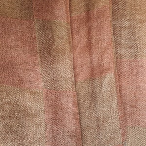 Op de afbeelding: Close-up van een geweven stof met een geruit patroon. De stof heeft afwisselende strepen van lichtbruin en stoffig roze. De textuur lijkt enigszins ruw, met zichtbare draden die een subtiel, getextureerd effect cre&euml;ren. De stof is gevouwen, waardoor zachte schaduwen ontstaan.
