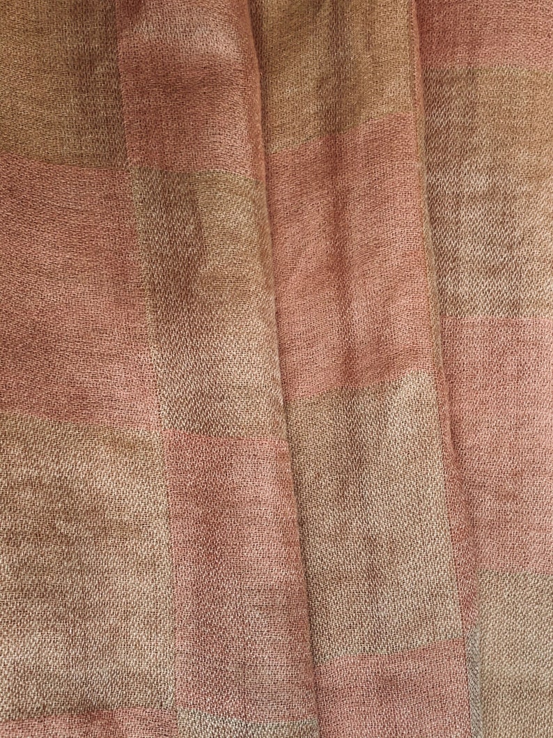 Op de afbeelding: Close-up van een geweven stof met een geruit patroon. De stof heeft afwisselende strepen van lichtbruin en stoffig roze. De textuur lijkt enigszins ruw, met zichtbare draden die een subtiel, getextureerd effect cre&euml;ren. De stof is gevouwen, waardoor zachte schaduwen ontstaan.