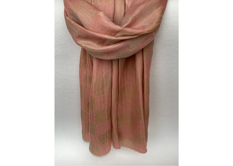 Op de afbeelding: Een zachte, geweven sjaal in roze en bruine tinten. De sjaal is gedrapeerd, wat de textuur en het patroon benadrukt. De stof lijkt lichtgewicht, met een subtiele glans. De randen zijn afgewerkt met een delicate franje.