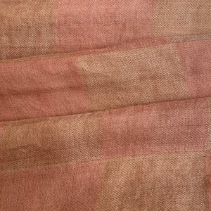 Op de afbeelding: Close-up van een textiel met een geweven patroon. De stof heeft afwisselende banden van gedempt roze en bruin, waardoor een subtiel dambordpatroon ontstaat. Het materiaal is zacht en lijkt gedrapeerd, met zichtbare plooien.