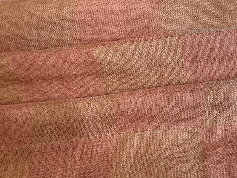 Op de afbeelding: Close-up van een textiel met een geweven patroon. De stof heeft afwisselende banden van gedempt roze en bruin, waardoor een subtiel dambordpatroon ontstaat. Het materiaal is zacht en lijkt gedrapeerd, met zichtbare plooien.