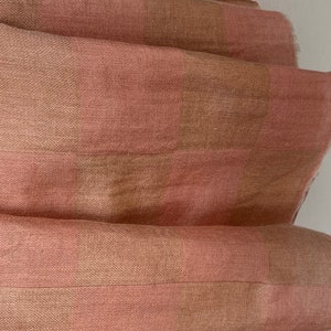 Op de afbeelding: Close-up van een gevouwen stof met een geruit patroon. De stof heeft afwisselende strepen van lichtroze en bruin. De textuur lijkt zacht en licht getextureerd, met zichtbare weefdetails. De algehele esthetiek is eenvoudig en elegant.