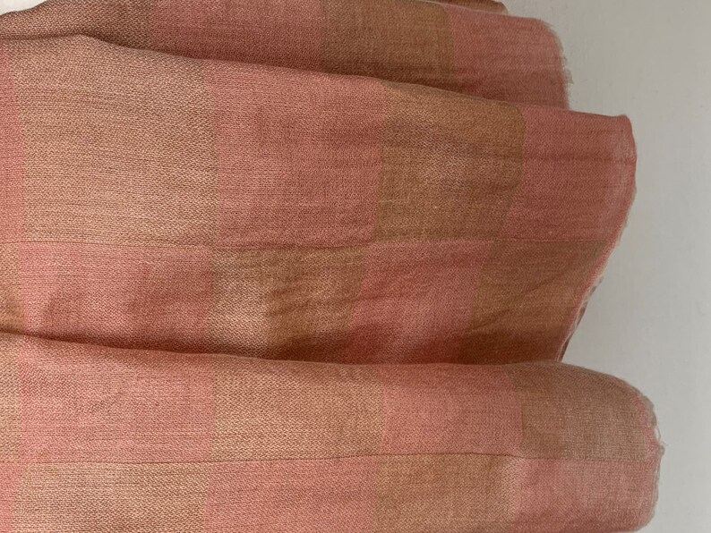 Op de afbeelding: Close-up van een gevouwen stof met een geruit patroon. De stof heeft afwisselende strepen van lichtroze en bruin. De textuur lijkt zacht en licht getextureerd, met zichtbare weefdetails. De algehele esthetiek is eenvoudig en elegant.