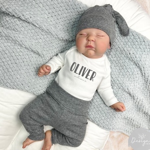 May include: A white baby onesie with the name "Oliver Williams" printed in gray. The onesie is worn by a baby wearing a gray beanie and gray pants.