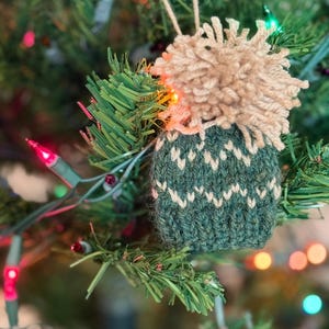 May include: A handmade Christmas tree ornament. The ornament features a green knitted base with a white pattern and a tan pom-pom top. The ornament is hanging on a Christmas tree with colorful lights.