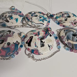 May include: Six white oval buttons with a colourful marbled design. Each button has two holes and is attached to a silver chain with pink and blue beads.