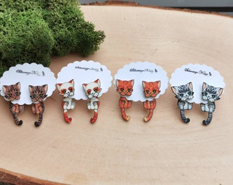 Könnte beinhalten: Vier Paar Ohrringe in Katzenform auf weißen Karten. Die Katzen sind in verschiedenen Farben und Mustern, darunter braunes Tabby, orangefarbenes Tabby, weiß mit orangefarbenen Flecken und grau mit weißen Streifen.