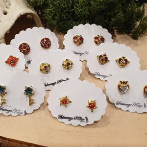 May include: Assortment of colorful enamel earrings on white cards. The earrings feature various designs, including hearts, stars, keys, and crowns. Each card has the text "Adoraway. K" printed on it.